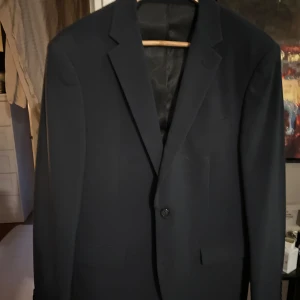 Marinblå kostym från J.Lindeberg - Snygg marinblå kostym från J.Lindeberg  med klassisk kavaj och matchande kostymbyxor. Kavajen har smala slag, enkelknäppning och diskreta fickor. Perfekt för dig som vill ha en stilren och tidlös look. Materialet är polyesterblandning för en skön känsla. Använd 1 gång förra året och kemtvättad efter så en är precis som ny. Köpt för 3900