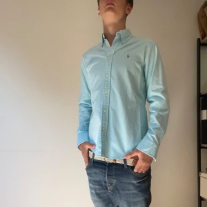 Turkos skjorta från Ralph Lauren - Snygg turkos skjorta från Ralph Lauren med klassisk button-down krage och broderad logga på bröstet. Skjortan har långa ärmar, knappar framtill och är tillverkad i mjuk bomull. Perfekt för en clean och stilren look. Modellen på bilden är 174 och väger 64kg.