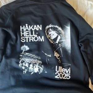Svart Håkan Hellström hoodie Ullevi 2025 - Svart hoodie med vit Håkan Hellström-tryck fram och stort konsertmotiv på ryggen med Ullevi 2025. Mjuk insida, huva med snörning och känguruficka. Perfekt för dig som älskar svensk pop och vill ha en snygg merchtröja.