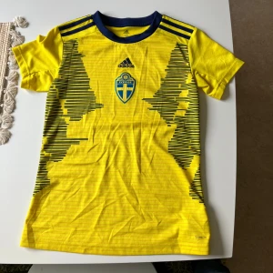 Sverige fotbollströja Adidas XS - Gul Sverige fotbollströja från Adidas i funktionsmaterial (Climalite). Tröjan har blå detaljer, blått landslagsmärke och Adidas-logga på bröstet samt svarta grafiska ränder på framsidan. Rund halsringning med blå kant och klassiska Adidas-ränder på axlarna.