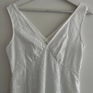 Vit långklänning med broderade blommor - Superfin vit långklänning med v-ringning och broderade blommor. Klänningen är ärmlös och har ett knytband i midjan för extra passform. Perfekt för sommarens alla tillfällen och ger en fräsch och romantisk vibe.