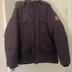 Mörkblå dunjacka Canada Goose XL - Säljer en mörkblå Canada Goose Constable dunjacka i storlek XL. Jackan har flera stora fickor, huva och klassisk Canada Goose-logga på ärmen. Perfekt för kalla dagar med sin rejäla och värmande design. Stängs med dragkedja och har en clean, stilren look.