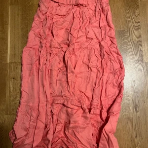 Lång rosa kjol från Rainbow - Säljer en lång, flowy kjol från Rainbow i en snygg rosa färg. Kjolen har resår i midjan och flera volangliknande partier som ger en cool och avslappnad vibe. Perfekt för dig som gillar färg och vill sticka ut lite extra i sommar.