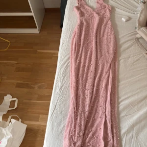 Rosa långklänning med pärlor - Supersnygg rosa långklänning med v-ringning och pärldetaljer över hela klänningen. Klänningen har smala axelband och en slits på sidan, perfekt för bal eller fest. Materialet känns lyxigt och skimrande.
