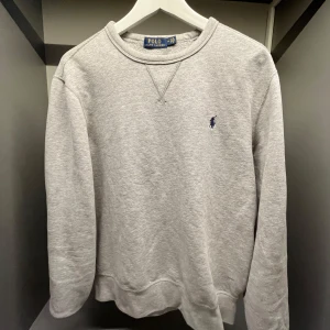 Grå sweatshirt från Polo Ralph Lauren - Klassisk grå sweatshirt från Polo Ralph Lauren med rund halsringning och broderad marinblå logga på bröstet. Tröjan har långa ärmar och ribbade muddar vid ärmslut och nederkant. Perfekt för en chill och stilren look.