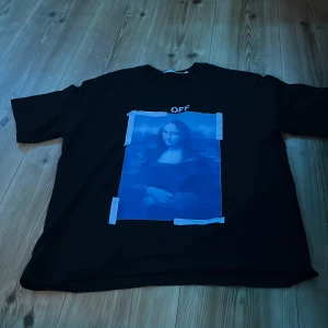 Off-White svart t-shirt Mona Lisa print - Svart t-shirt från Off-White med stort blått Mona Lisa-print framtill och klassiska vita pilar på ryggen. Oversized passform och korta ärmar. Tryckt Off-White-logga ovanför motivet. Perfekt statement-plagg för dig som gillar streetwear.