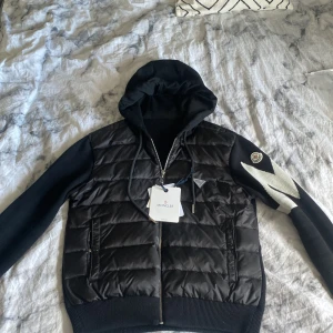 Svart Moncler cardigan med huva - Svart cardigan från Moncler med quiltad front i syntetmaterial och stickade ärmar. Jackan har huva, dragkedja och en vit zigzag-detalj på ena ärmen samt Moncler-logga. Perfekt för dig som vill ha en stilren och sportig look.