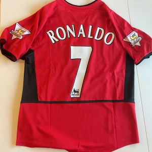 Manchester United Ronaldo 7 Nike tröja - Klassisk Manchester United replika matchtröja med Ronaldo 7 på ryggen, Premier League-märken på ärmarna och Vodafone som sponsor. Röd med svarta detaljer, kortärmad och tillverkad i lätt polyester. Perfekt för dig som älskar fotboll och vill ha en ikonisk retrotröja. Endast handtvätt!