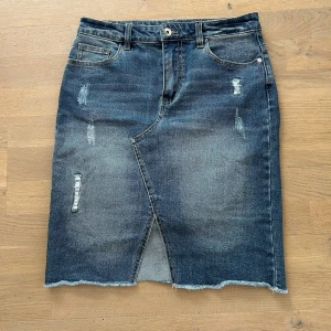 Mörkblå jeanskjol med slits - Snygg mörkblå jeanskjol med slitna detaljer, rå kant nertill och en cool slits framtill. Klassisk femficksmodell med bälteshällor och bakfickor. Perfekt för dig som gillar en avslappnad och trendig stil.