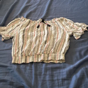 Randig offshoulder blus från H&M - Supersöt offshoulder blus från H&M i vitt med svarta och rosa ränder. Blusen har korta ärmar med volangkant, smockad nederkant och en svart knyt-detalj framtill. Perfekt för en chill sommardag eller när du vill ha en boho-vibe.