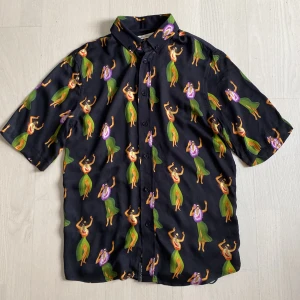 Svart skjorta med hawaiimönster The Blue Uniform strl S - Cool svart skjorta från The Blue Uniform med färgglatt hawaiimönster av dansande figurer i grönt, lila och orange. Skjortan har korta ärmar, klassisk krage och knäppning framtill. Tillverkad i mjuk viskos som känns skön mot huden.