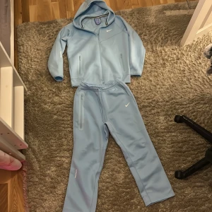 Nike NOCTA ljusblå sett  - Säljer en stilren ljusblå vindjacka från Nike NOCTA med huva och dragkedja. Jackan har två fickor med dragkedja, NOCTA-tryck på ärmen och klassisk Nike Swoosh på bröstet. Perfekt för dig som gillar sportig och clean stil. helt ny pris kan diskuteras