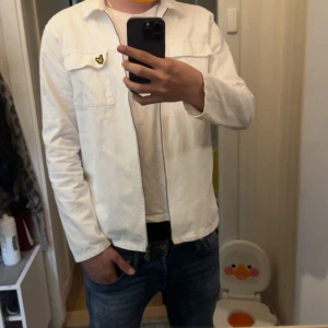 Vit overshirt från Lyle & Scott - Säljer en stilren vit overshirt från Lyle & Scott i storlek S. Skjortan har dragkedja framtill, två bröstfickor och den klassiska gula loggan på vänster sida. Perfekt att slänga över en t-shirt för en clean look.