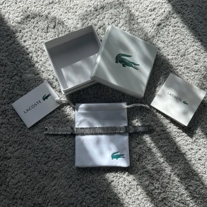 Lacoste Metropole armband i silver - Snyggt Metropole armband från Lacoste i silverfärgad metall med stilren, fyrkantig länkdesign. Kommer med original ask, tygpåse och certifikat. Perfekt accessoar för dig som gillar klassisk och modern stil med en touch av sportig känsla.