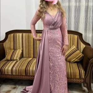 Lång lila festklänning med spets - Säljer en elegant långklänning i lila med glittriga spetsdetaljer och omlott-effekt över bysten. Klänningen har långa ärmar och ett vackert släp i satinliknande material. Perfekt för bal eller festliga tillfällen.