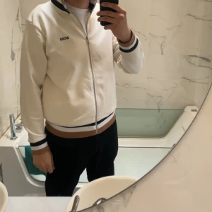 Vit tröja från BOSS - Snygg vit tröja  från BOSS med svarta och bruna detaljer vid muddar och krage. Jackan har dragkedja framtill och en stilren design med logga på bröstet. Perfekt för dig som gillar en clean och sportig look.
