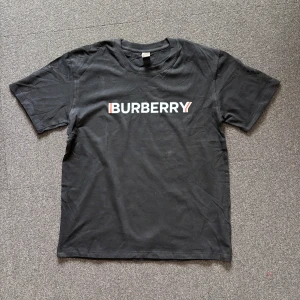 Svart Burberry t-shirt - Svart t-shirt från Burberry med klassisk vit logga framtill och röda detaljer. T-shirten har rund halsringning och korta ärmar. Tillverkad i mjuk bomull för en skön känsla och enkel stil. Den kan passa både storlek M och S.