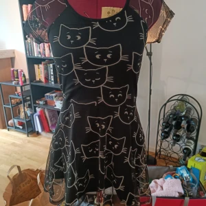 Svart meshklänning med kattmönster - Supersöt svart kortklänning i mesh med vita kattansikten över hela tyget. Klänningen har korta ärmar i transparent mesh och en lätt utställd kjol. Perfekt för dig som älskar unika och lekfulla plagg med djurmotiv. Finns en liten rosa fläck. Se bild!!
