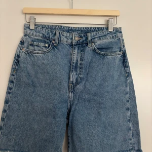 Blå loose jeansshorts från &Denim - Snygga blå jeansshorts från &Denim med loose fit och hög midja. Klassisk femficksmodell, råa kanter nertill och dragkedja framtill. Perfekta för sommaren och ger en avslappnad vibe.