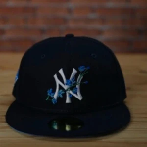 Svart New York Yankees keps med blommor - Snygg svart keps från New York Yankees med broderade blå blommor runt det klassiska NY-loggan framtill. Platt skärm och cool streetstil, perfekt för att lyfta din outfit med en unik detalj. Strl 56.8 är prutbar vid snabbaffär 