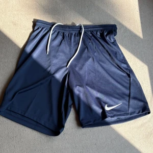Marinblå Nike shorts med snörning - Snygga marinblå shorts från Nike med vit snörning i midjan och klassisk Swoosh-logga på benet. Tillverkade i 100% polyester för en lätt och sportig känsla. Perfekta för träning eller chill dagar när du vill ha maximal rörelsefrihet.