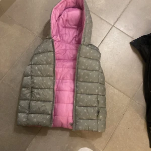 Vändbar rosa och grå dunväst med luva - Vändbar dunväst med luva, ena sidan ljusgrön med små blommor och andra sidan ljusrosa. Västen är quiltad och har dragkedja framtill. Perfekt för dig som vill kunna variera din stil snabbt. Snygg och mysig att bära över hoodie eller tröja.