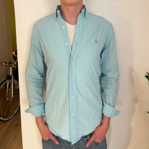 Ljusblå skjorta från Polo Ralph Lauren - Snygg ljusblå skjorta från Polo Ralph Lauren i slim fit-modell. Klassisk button-down krage, broderad logga i lila på bröstet och långa ärmar. Skjortan är i bomull och har knappar framtill. Perfekt för en clean och stilren look.