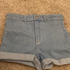 Ljusblå jeansshorts från &Denim - Säljer ett par ljusblå jeansshorts från &Denim med klassisk femficksdesign och uppvikta benslut. Shortsen har bälteshällor och knapp framtill. Perfekta för varma dagar och enkel att matcha med olika toppar.