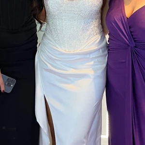 Vit långklänning med slits och spetsliv - Elegant vit långklänning med figurnära passform, spetsdetaljer på livet och markerad slits framtill. Perfekt för bal eller fest. Axelbandslös design som ger en stilren och lyxig känsla.