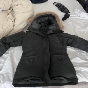 Svart dunjacka med pälskant från Canada Goose - Säljer en svart dunjacka från Canada Goose med huva och pälskant. Jackan har långa ärmar, knappar och dragkedja framtill samt två fickor med lock. Perfekt för kalla dagar och riktigt varm. Rätt fint skick, lite gammal bara