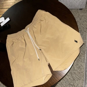 Beige M badshorts från Polo Ralph Lauren - Snygga beige bad shorts från Polo Ralph Lauren med vit snörning i midjan och klassisk broderad logga på benet. Perfekta för sommaren och har en avslappnad passform i M - Medium