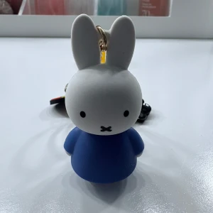 Miffy nyckelring med blå klänning - Söt nyckelring med Miffy-figur i blå klänning och vit kropp. Nyckelringen har ett gult band med texten 'miffy' i rött och flera små dekorationer. Perfekt som accessoar till väskan eller nycklarna.
