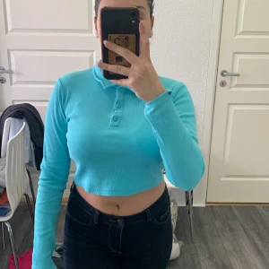Turkos croppad långärmad pikétröja från FB Sister - Säljer en turkos croppad pikétröja från FB Sister i ribbat material. Tröjan har långa ärmar, klassisk krage och knappar framtill. Perfekt för dig som gillar färg och vill ha en bekväm men snygg topp.