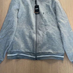 Grå zip-tröja från BOSS - Snygg grå jacka från BOSS med dragkedja framtill och broderad logga på bröstet. Jackan har långa ärmar och ribbade muddar med vita ränder vid ärmslut och nederkant. Perfekt för en stilren och avslappnad look.