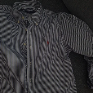 Blåvit randig skjorta från Ralph Lauren - Klassisk blåvit randig skjorta från Ralph Lauren med button-down krage och broderad röd logga på bröstet. Skjortan har långa ärmar och knäppning framtill. Perfekt för en stilren look.