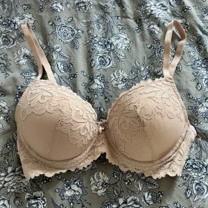 Beige spets-bh med rosett - Oanvänd. Säljer en beige bh med vacker spets och liten rosett framtill. Bh:n har vadderade kupor och justerbara axelband. Perfekt för dig som vill ha något både snyggt och bekvämt.
