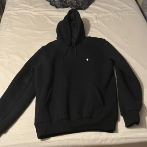Svart hoodie från Ralph Lauren - Säljer en svart hoodie från Ralph Lauren med klassisk broderad logga på bröstet. Tröjan har huva med dragsko och en stor magficka. Perfekt för en avslappnad och stilren look.