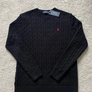 Kabelstickad tröja från Ralph Lauren - Säljer en svart kabelstickad tröja från Ralph Lauren med rund halsringning och röd broderad logga på bröstet. Tröjan har långa ärmar och ribbade muddar vid ärmslut och nederkant. Perfekt för en stilren look. (Lapp finns men sitter ej på. Tröjan är bara använd någon enstaka gång).