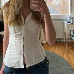 Vit korsett-topp med knappar - Säljer en vit korsett-topp med knappar framtill och markerad midja. Toppen har v-ringning och är ärmlös, vilket ger en snygg och modern siluett. Perfekt att styla med jeans eller kjol för en trendig look.