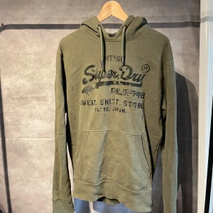 Superdry Hoodie - Säljer en olivgrön hoodie från Superdry med tryckt logga och text på bröstet. Tröjan har huva med dragsko och en stor magficka framtill. Perfekt för en avslappnad och cool stil.