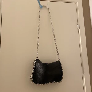 Svart axelväska med kedja - Säljer en svart axelväska med grov silverfärgad kedja som axelrem och dekor runt kanterna. Väskan har en mjuk, lätt glansig yta och är tillverkad i skinnimitation. Aldrig användt💕