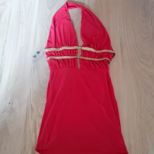 Röd halterneckklänning med gulddetaljer - Snygg röd halterneckklänning med guldiga band i midjan och dekorativt spänne framtill. Klänningen har djup ringning och knyts i nacken. Perfekt för dig som vill sticka ut med färg och detaljer. Använd en kväll. Tyvärr så fick den trådsläpp på banden då. Dyrt nypris. Tyvärr finns inga lappar inuti. Jag minns inte vars jag köpte den. Men den är 100% säkert inte ifrån temu, shein eller liknande. Perfekt till fest, party. Så snygg rygg 