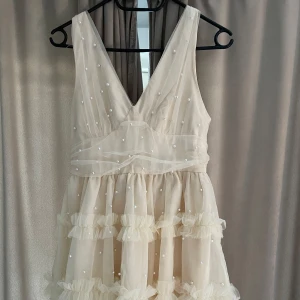 Studentklänning med volanger och pärldetaljer - Supersöt vit kortklänning med djup v-ringning och volanger i flera lager. Klänningen har pärldetaljer över hela tyget och är ärmlös. Perfekt för sommarens speciella tillfällen!