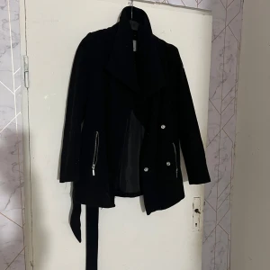 Svart kappa från Vero Moda - Stilren svart kappa från Vero Moda med bred krage, knappar och dragkedjor vid ärmarna. Jackan har ett bälte i midjan och är helsvart, perfekt för dig som gillar en klassisk look.