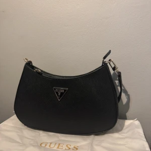 Svart axelväska från Guess - Snygg svart axelväska från Guess med klassisk triangellogga i metall framtill. Väskan har justerbar axelrem och stilren design, perfekt för att lyfta vilken outfit som helst. Materialet är strukturerat syntet och väskan har dragkedja upptill.