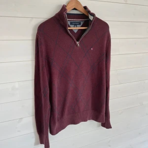 Vinröd halvzip tröja från Tommy Hilfiger - Vinröd stickad tröja från Tommy Hilfiger med halv dragkedja och diskret rutmönster i blått. Tröjan har lång ärm, ribbade muddar och liten logga på bröstet. Perfekt för lager-på-lager och snygg till jeans.