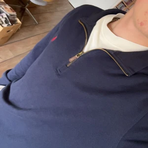 Mörkblå tröja med dragkedja från Polo Ralph Lauren - Säljer en mörkblå långärmad tröja från Polo Ralph Lauren med halv dragkedja och klassisk röd logga på bröstet. Perfekt för en stilren och avslappnad look. Snygg krage och mjukt material. 