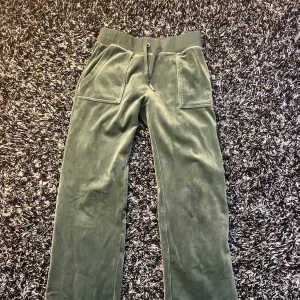 Gröna velourbyxor från Juicy Couture - Mörkgröna velourbyxor från Juicy Couture med bred resår i midjan och snörning. Byxorna har raka ben, stora fickor fram och bak samt en broderad JC-logga på ena benet. Perfekta för en avslappnad och trendig look.