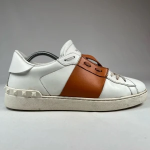 Valentino Garavani sneakers med bruna detaljer - Säljer ett par vita sneakers från Valentino Garavani med breda bruna läderdetaljer över snörningen och nitar på hälen. Skorna har rund tå, platt sula och klassisk snörning. Perfekta för dig som gillar stilrena men ändå unika sneakers.