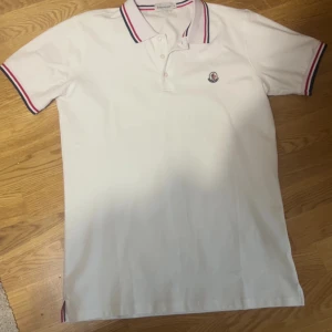 Vit pikétröja från Moncler - Säljer en klassisk vit pikétröja från Moncler med broderad logga på bröstet. Tröjan har korta ärmar och krage med röd och marinblå rand, samt matchande detaljer vid ärmslut. Perfekt för en stilren och sportig look. Qr kod funkar ej